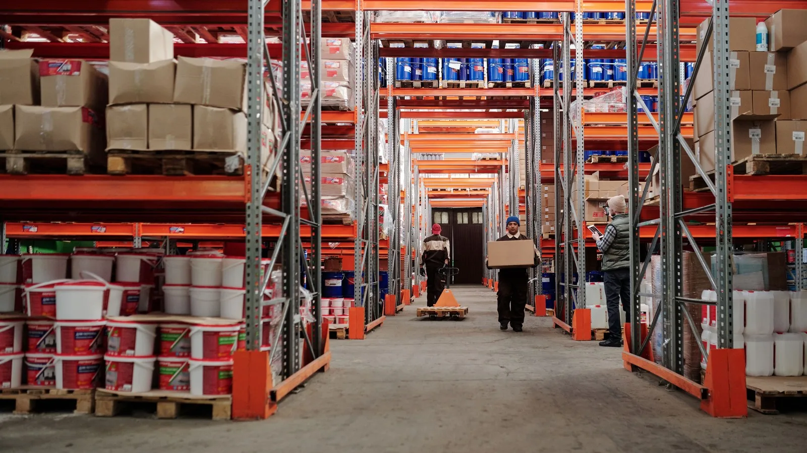 Warehouse workers moving boxes and a pallet jack down a busy aisle between tall racking